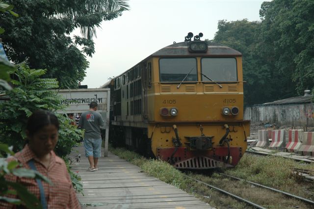 The Thai railway network runs through the main city of Bangkok, in many places. The diesel powered passenger train has come to a halt in what appears to be a rural station, but, metres behind the photographer, is a very busy city intersection